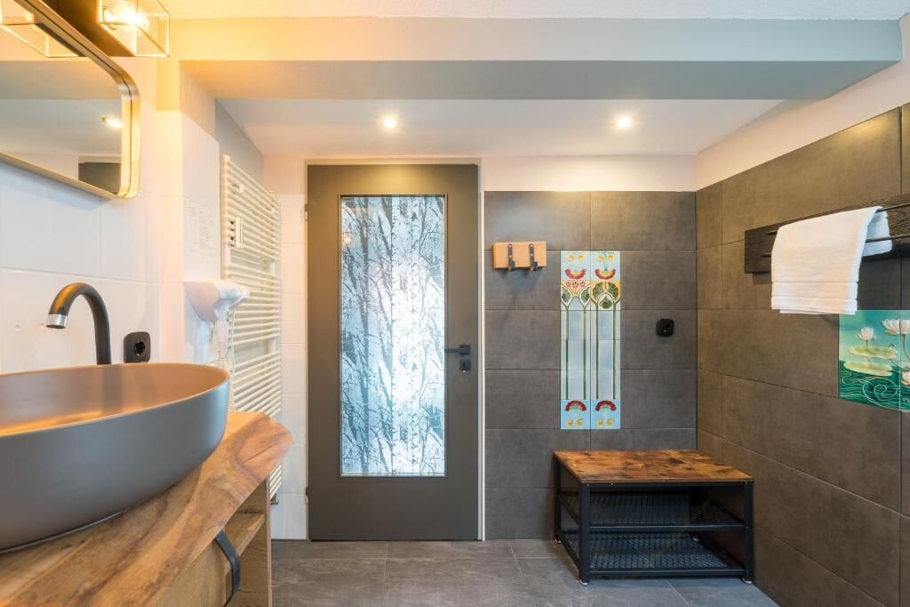 Ein modernes Badezimmer mit einem großzügigen Aufgussbecken, einer Holzbank, einer Tür mit Milchglasfüllung und dekorativen Fliesen.