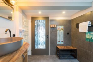 A modern bathroom with a large vessel sink, a wooden bench, a door with a frosted glass panel, and decorative tiles. - Design Apartments - Remise Blumberg (Potsdam)