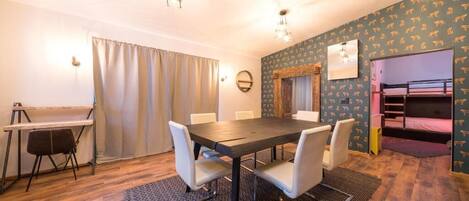 A room with a wooden dining table, white chairs, a bunk bed, a desk, and a ceiling fan.