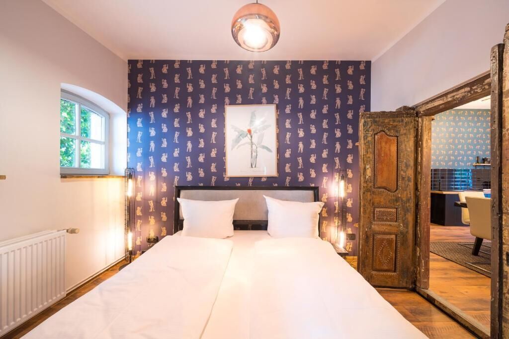 A bedroom with a large bed, a window, a door leading to another room, and a wall with a patterned wallpaper.
