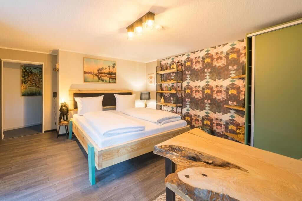 Ein Schlafzimmer mit einem hölzernen Bett, einem Nachttisch, einer Lampe, einer Wand mit geometrischem Muster und einem Landschaftsbild.