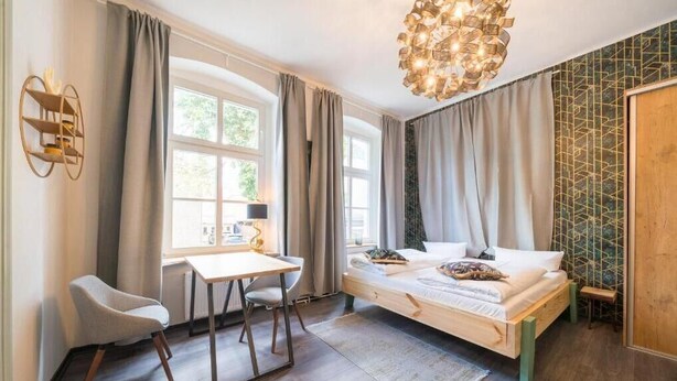 A modern bedroom with a wooden bed, a desk, and a chair. There is a window with curtains, a chandelier, and a geometric-patterned wall. - Design Apartments - Remise Blumberg (Potsdam)