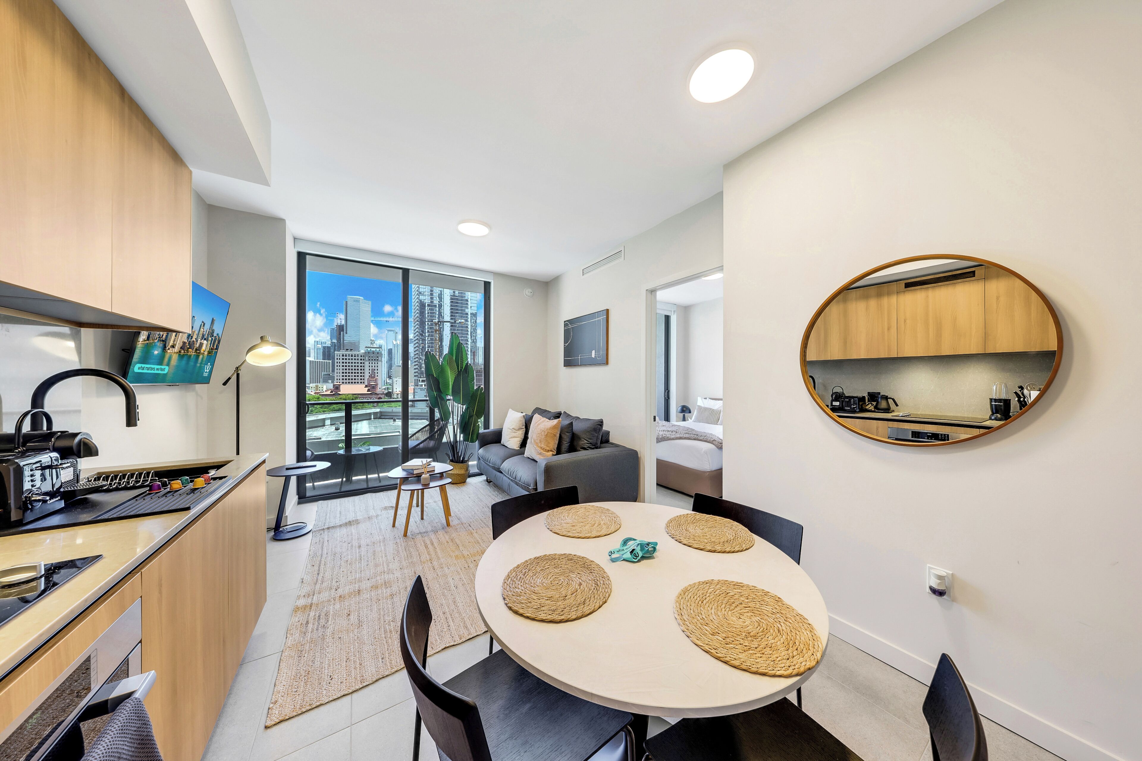 A modern kitchen with a built-in espresso machine, a dining area with a round table and black chairs, and a living space with a sofa and a large window overlooking a cityscape.