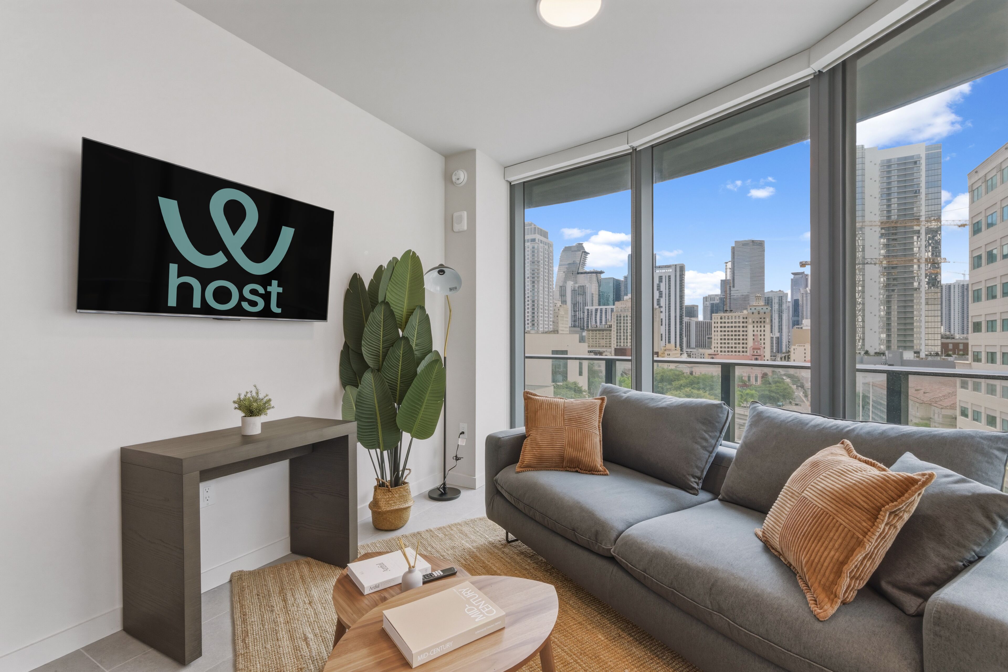 A modern living room with a city view, a TV mounted on the wall, a grey sofa, and a console table with a plant.