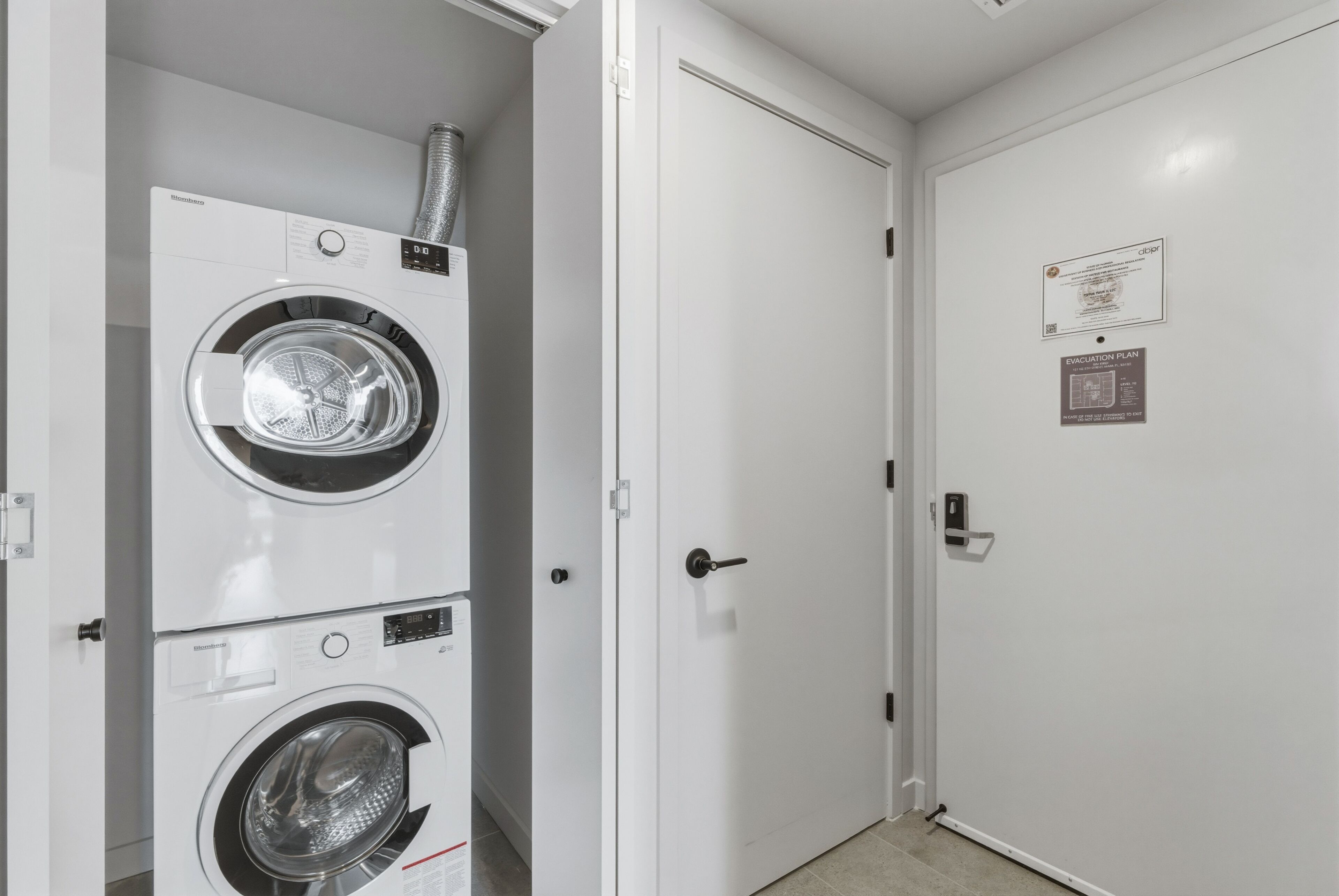 A laundry room with stacked washers and dryers, a white door with a fire safety sign, and a tiled floor.