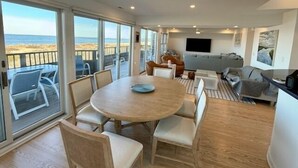 Un salón amplio con una gran mesa redonda, asientos cómodos, un televisor y un balcón con vista a la playa.