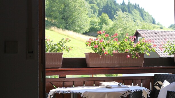 En balkong med träräcke, ett bord med ett vitt bordsduksöverdrag och krukväxter med rosa blommor.
