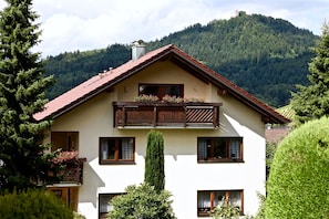 Una casa a due piani con tetto rosso, balcone in legno e montagne sullo sfondo.