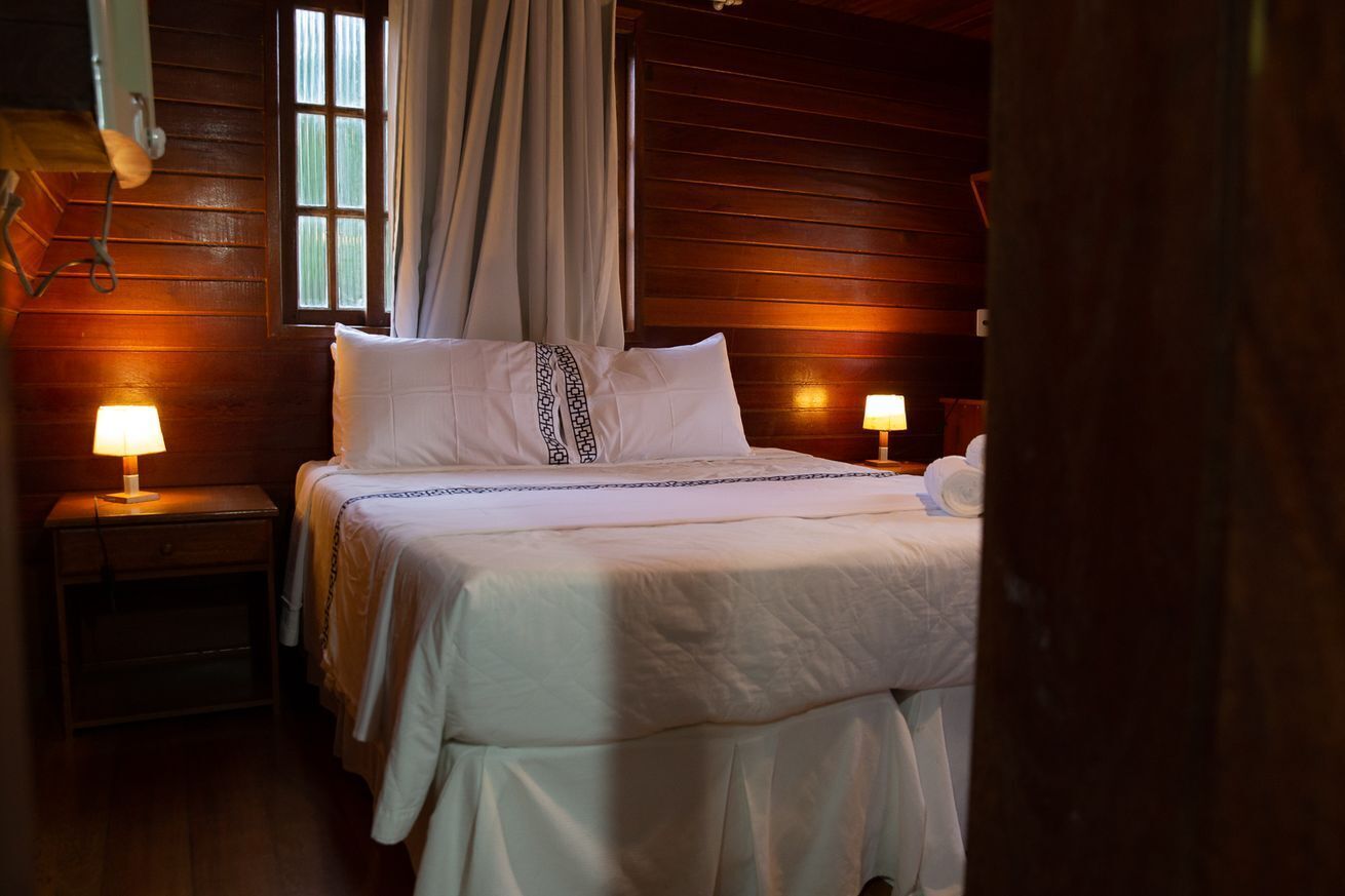 A cozy bedroom with a wooden cabin-style interior, featuring a large bed with white bedding and a patterned pillow, two bedside lamps, and a window with curtains.