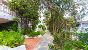 A stone pathway flanked by lush greenery and flowering plants, leading to a white building with a balcony. - Beach Access Apt in Playa Real | Pool & Terrace - Pass the Keys (Marbella)