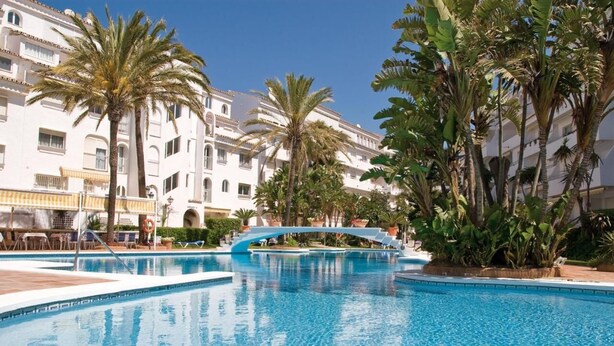 A swimming pool with a blue bridge, surrounded by palm trees and white buildings with balconies. - Beach Access Apt in Playa Real | Pool & Terrace - Pass the Keys (Marbella)