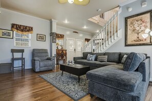 Ein Wohnzimmer mit grauem Ecksofa, einem kleinen grauen Sessel, einem Couchtisch und einem gemusterten Teppich. Eine Treppe mit weißen Geländern und ein gerahmtes Blumenbild an der Wand sind ebenfalls zu sehen.