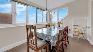 A dining area with a table covered by a sailboat-themed tablecloth, surrounded by wooden chairs with cushions. - Jarrahdale Beach View Apartment - Pass the Keys (Herne Bay)