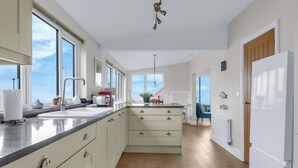 A modern kitchen with a large island, ample counter space, and a view of the sky through large windows. - Jarrahdale Beach View Apartment - Pass the Keys (Herne Bay)