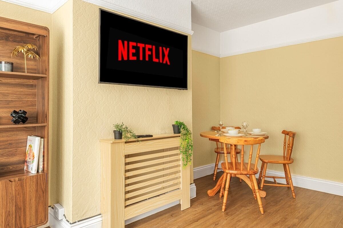 Ett rum med ett TV-bord i trä, ett matbord med stolar och en väggmonterad TV som visar ”NETFLIX”.