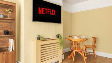 Ett rum med ett TV-bord i trä, ett matbord med stolar och en väggmonterad TV som visar ”NETFLIX”.