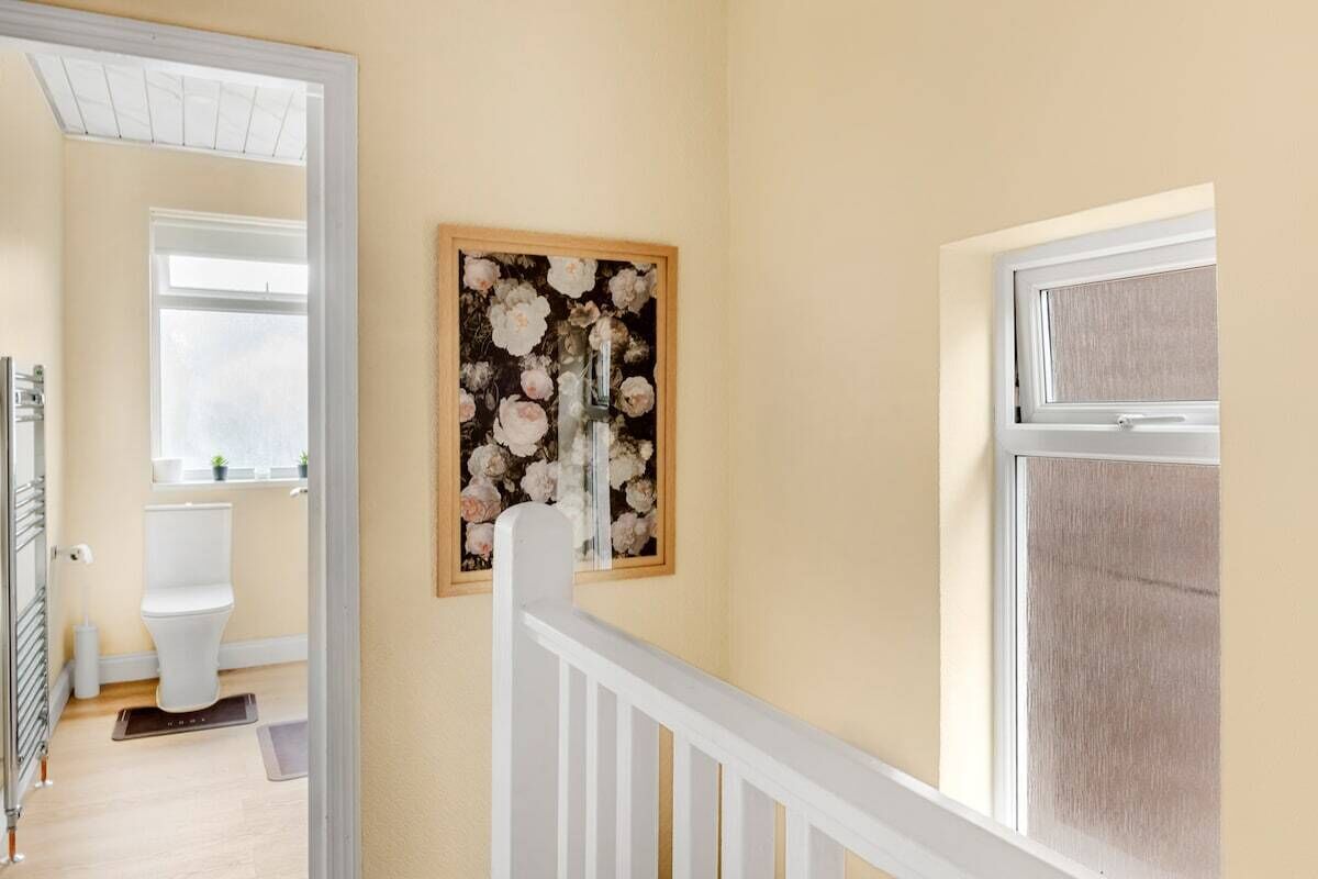 A hallway with a white railing, a framed floral artwork on the wall, and a view into a bathroom with a toilet and a window.
