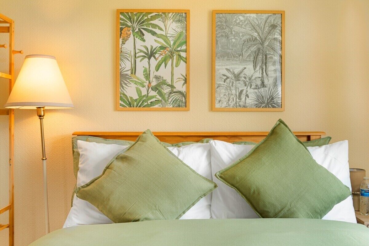 A bed with green pillows, a bedside lamp, and two framed botanical prints on the wall.