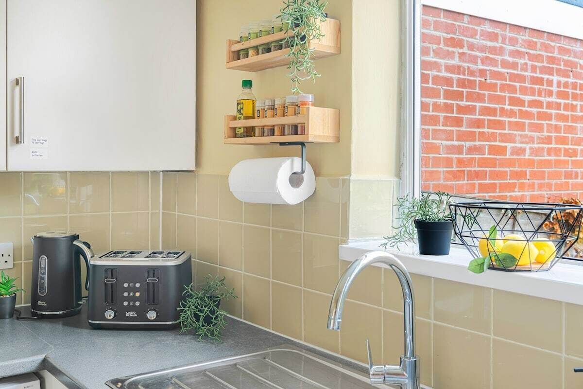 Et køkken med indbygget toaster, elkedel og vask med armatur. Over vasken er der træhylder med krukker og en plante.