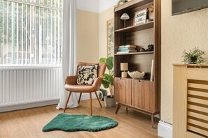 A room with a wooden floor, a leather chair with a floral pillow, a green rug, a wooden cabinet with shelves, and a window with blinds. - Rosewick Villa wt Office, Garden & Free Parking (Nottingham)