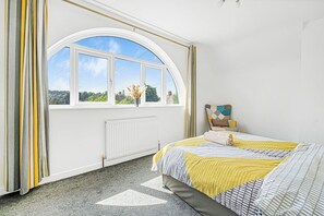 Une chambre avec une grande fenêtre en arc de cercle, un lit recouvert d’une courtepointe à motifs et un radiateur placé sous la fenêtre.