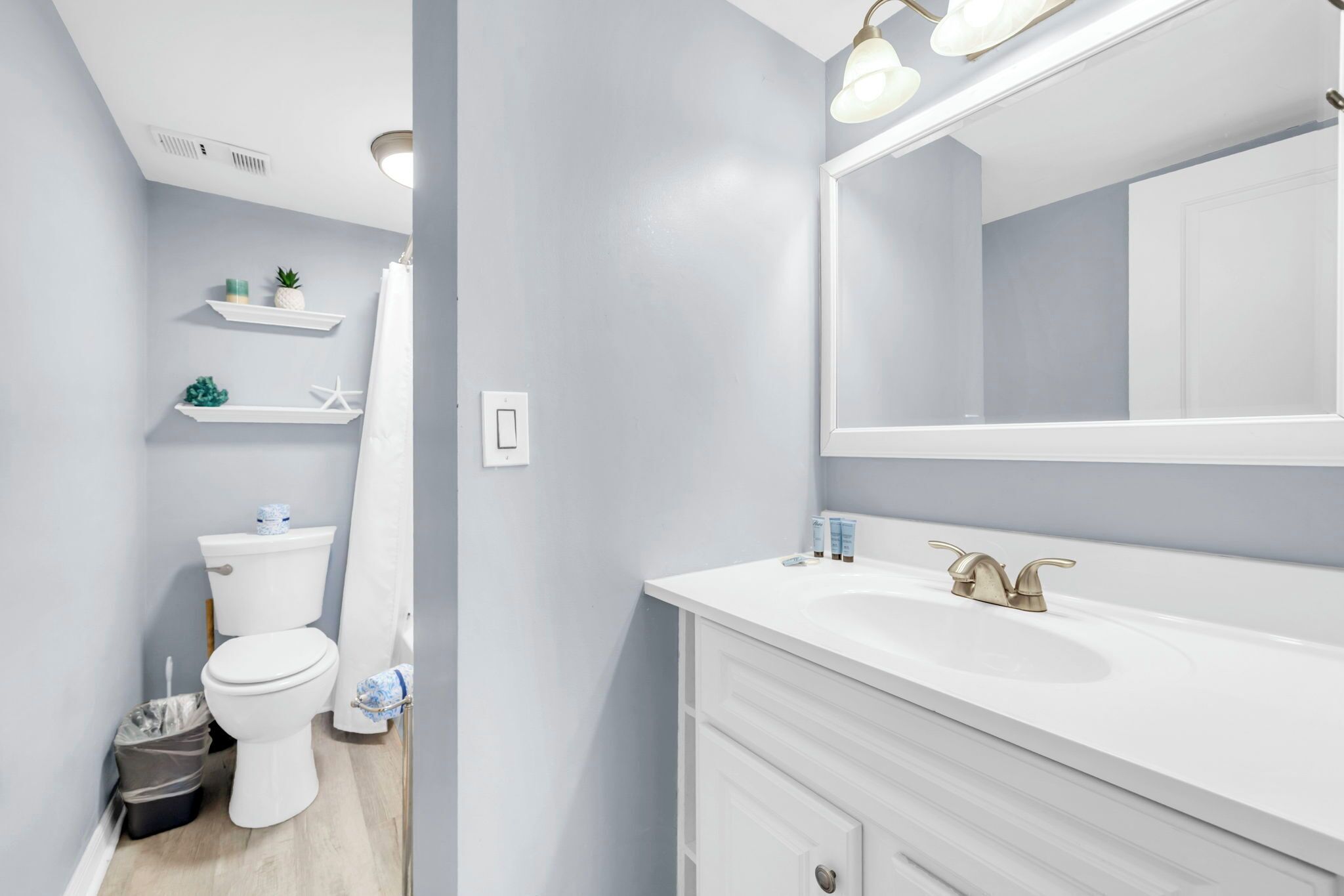 A bathroom with a white toilet, a sink with a gold faucet, and a mirror above the sink. Shelves with toiletries are visible on the wall.
