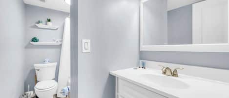 A bathroom with a white toilet, a sink with a gold faucet, and a mirror above the sink. Shelves with toiletries are visible on the wall.
