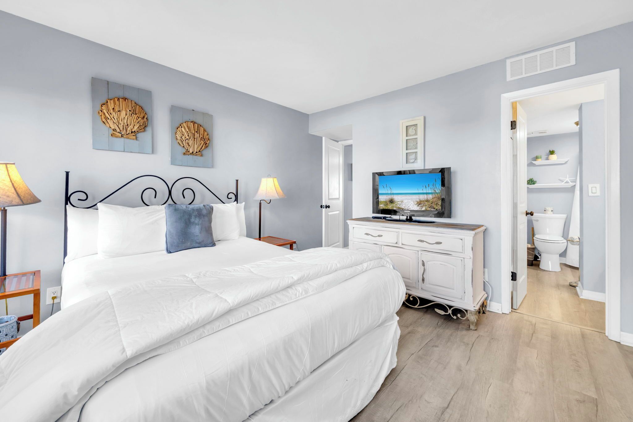 A bedroom with a large bed, a TV, and a dresser. There is a bathroom visible through an open door.