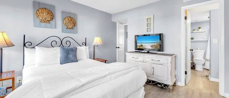 A bedroom with a large bed, a TV, and a dresser. There is a bathroom visible through an open door.