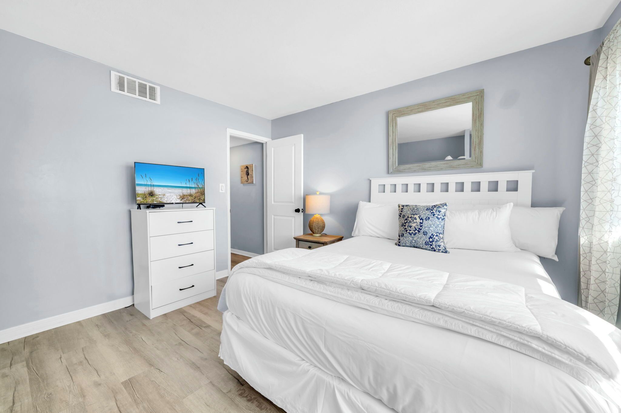 A bedroom with a bed, a TV, a nightstand with a lamp, and a door leading to another room.