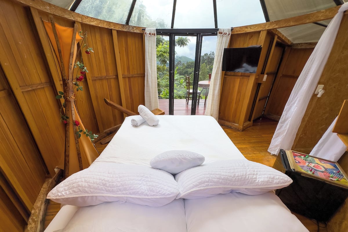 A room with a bed, wooden walls, a TV, and a view of greenery outside.
