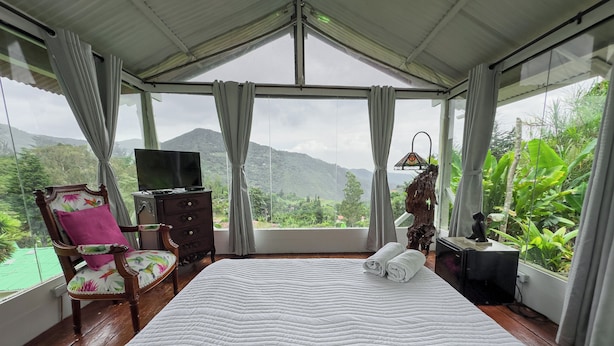 A bedroom with a large bed, a TV, a dresser, a chair, and a view of a mountainous landscape through large windows.