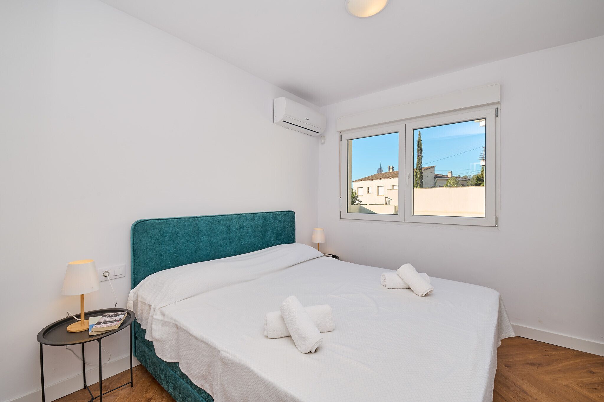 Una cama bien hecha con sábanas blancas y dos toallas enrolladas, un cabecero color turquesa, una lámpara de noche y una ventana con vistas a edificios.