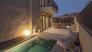 A modern outdoor pool area with a lounge chair and a dining table, surrounded by a low fence and a building with a balcony. - Villa Villagio Rosso (Kokkino Chorio)
