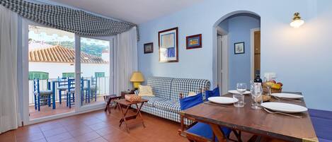 A dining area with a wooden table set for two, a blue sofa, and a view through a glass door to an outdoor terrace.