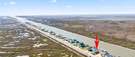 An aerial view of a coastal area with a road lined with buildings, a river, and marshy terrain.