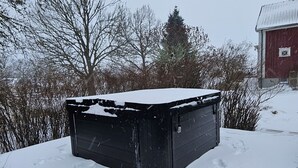 A snow-covered outdoor area with a black object on a wooden platform, surrounded by trees and a red building in the background. - Apartment at country side. Spa facility and much more out in swedish woods. (säffle)