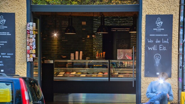 A bakery with a display case of baked goods, a sign with text, and a person sitting outside. - Family Apartment in Berlin for 6 – Central & Quiet, Supermarket & Park Nearby (Berlin)