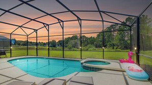 A covered swimming pool with lounge chairs and a hot tub, surrounded by a metal fence and a view of a grassy field. - Villa Maggiore on Watersong (Davenport)