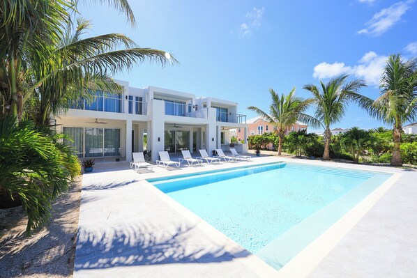 Une maison moderne à deux étages, dotée d’une piscine, entourée de palmiers et surplombée d’un ciel bleu azur.