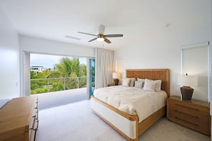 Une chambre moderne avec un grand lit, un ventilateur de plafond, une table de chevet avec une lampe et une vue sur un balcon agrémenté de plantes.