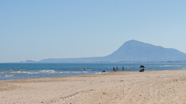 Uma praia com uma montanha ao fundo, algumas pessoas e um cachorro.