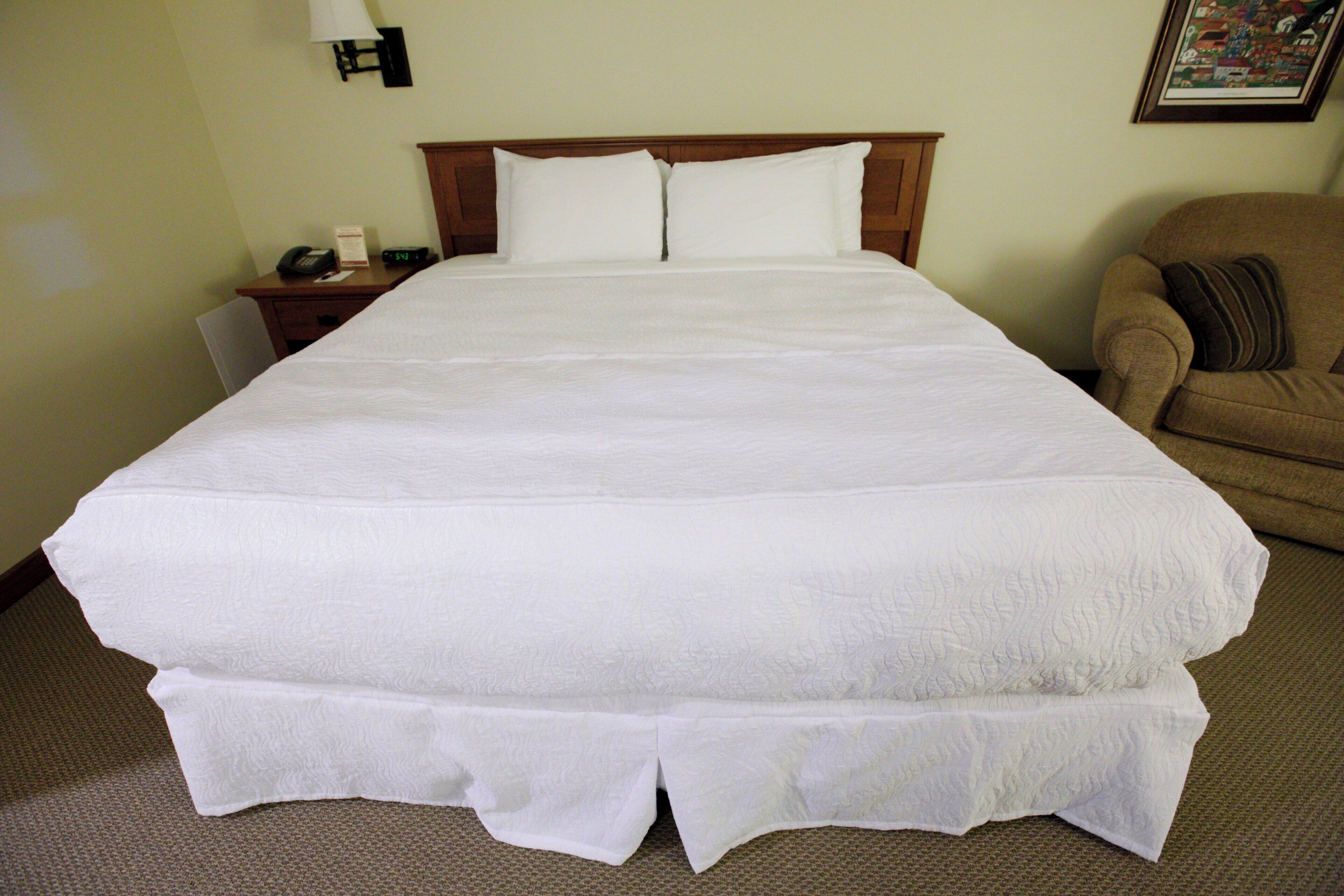 A large bed with white bedding, a wooden headboard, a nightstand with a lamp, and a chair with a cushion.