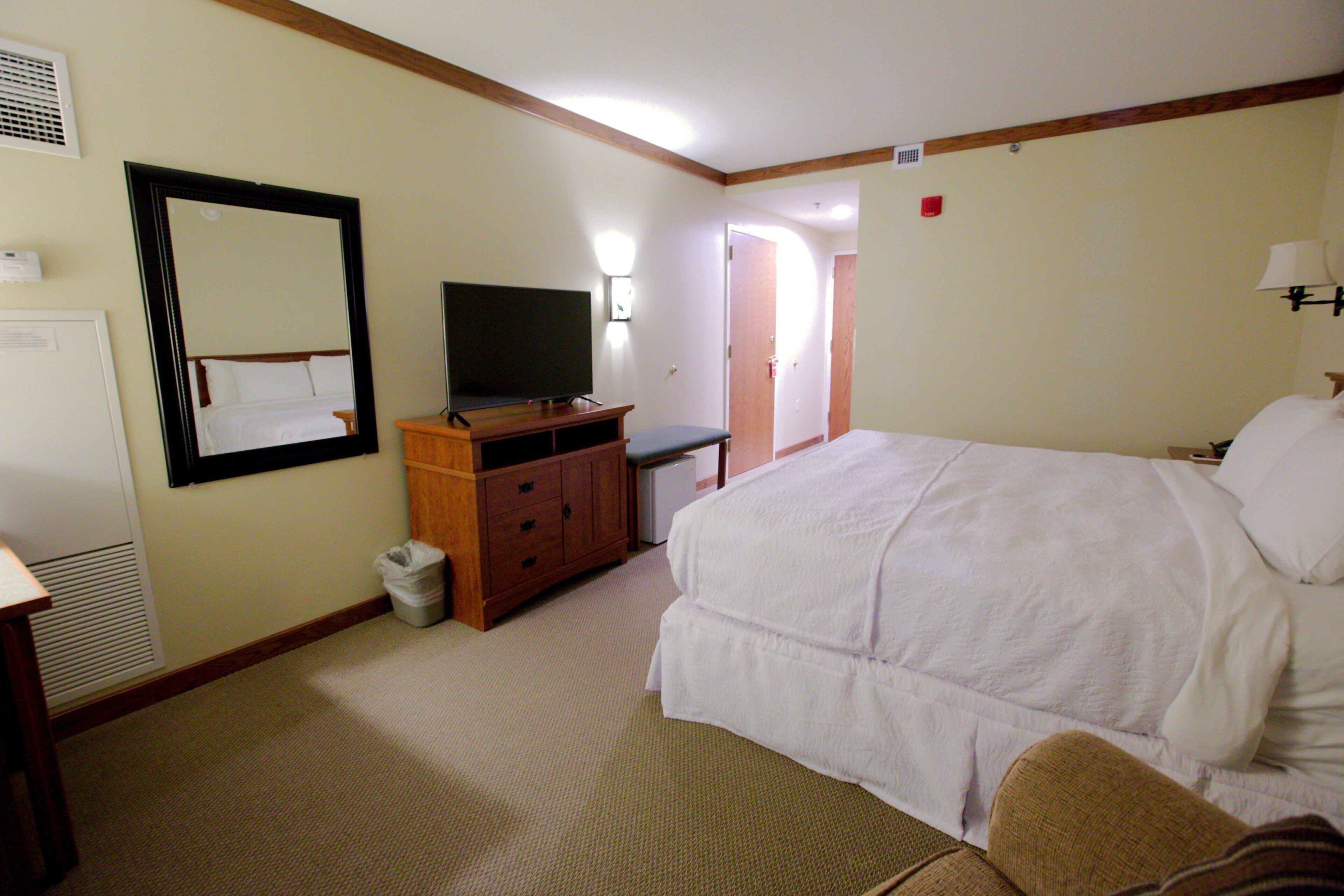 Ein Hotelzimmer mit einem Bett, einem Fernseher, einem Schminktisch, einem Spiegel und einer Tür zu einem weiteren Raum.
