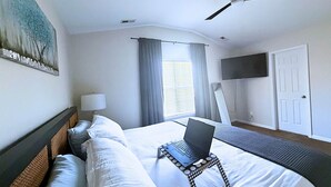 Ein Hotelzimmer mit einem Bett, einem auf einem Ständer stehenden Laptop, einem Deckenventilator, einem Fenster mit Vorhängen, einem Flachbildfernseher und einer Tür.