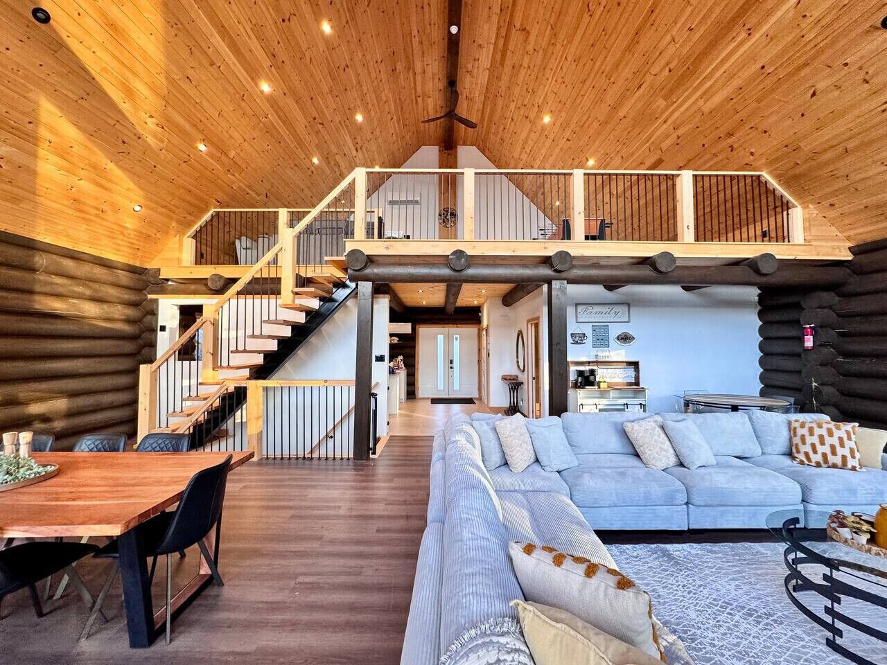 A spacious log cabin interior with a wooden staircase leading to a loft bedroom, a dining area, and a living room with a sofa and coffee table.