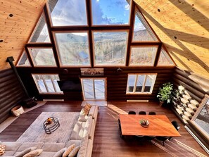 A cozy cabin interior with a wooden interior, a sofa, a dining table, and large windows offering a view of the forest. - Le Nørdéa | Spa | Chiens permis | Bois Rond | (Saint-Côme)