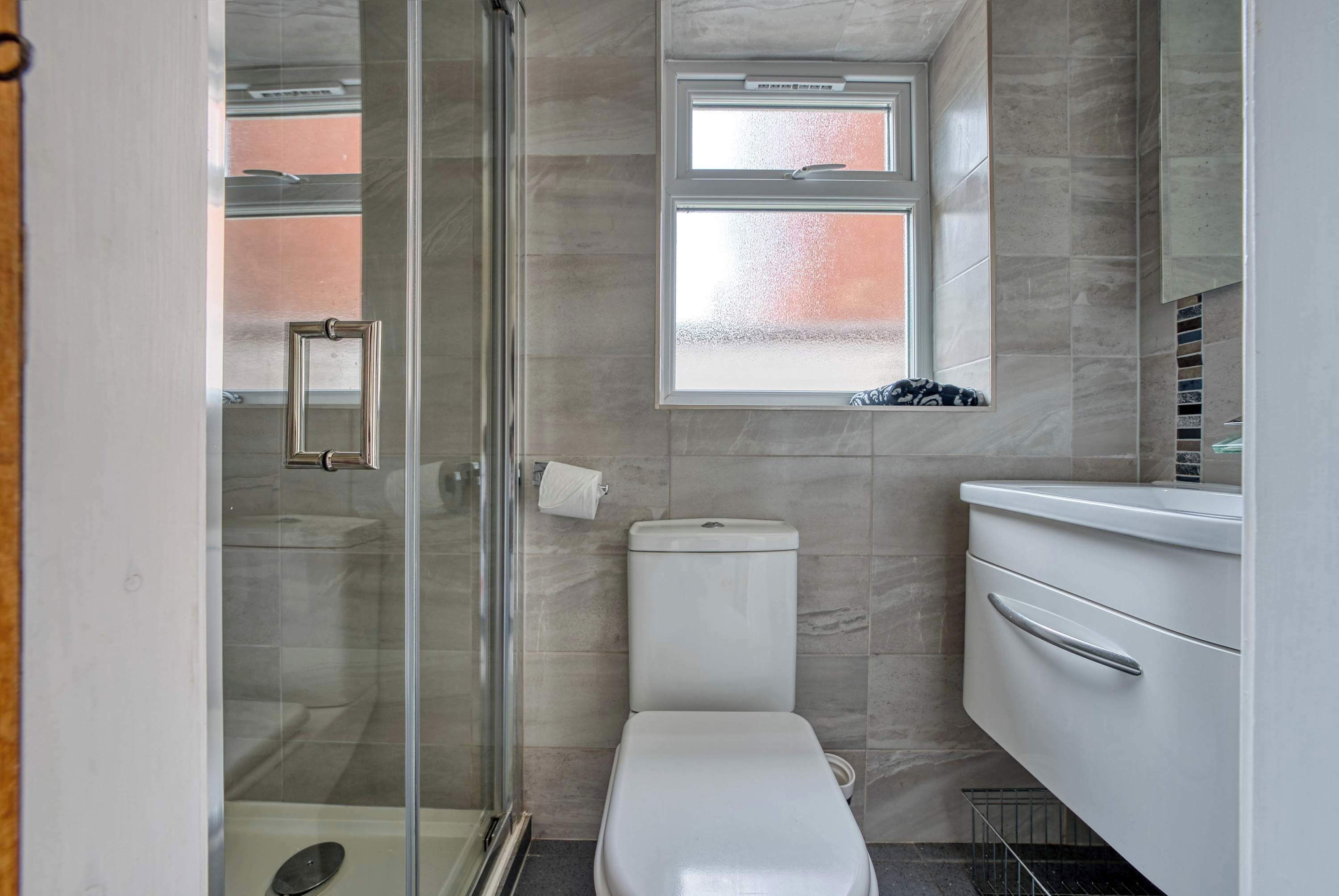 A bathroom with a glass shower enclosure, a toilet, a window, and a white sink with a mirror.
