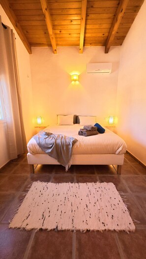 A bedroom with a bed, two bedside lamps, a window with curtains, and a wooden ceiling.