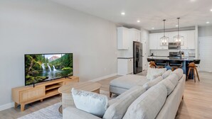 A modern living room with a large flat-screen TV, a grey sofa, wooden entertainment unit, and a kitchen area with white cabinets and a stainless steel refrigerator.